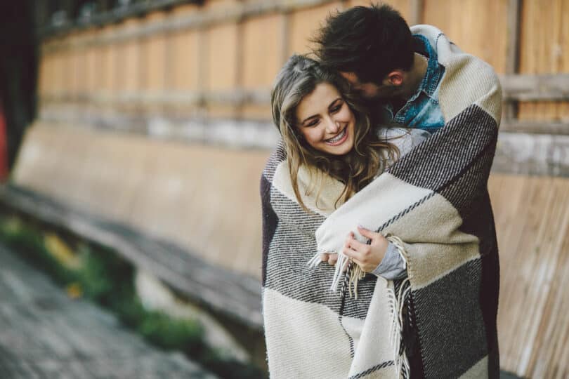 Casal enrolado em um cobertor no foco com mulher sorrindo e homem com cabeça apoiada no cabelo dela