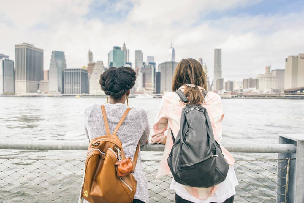 Duas mulheres, uma negra e uma branca, lado a lado, observando uma paisagem urbana de prédios, próximas à um rio