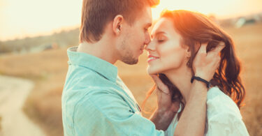 Casal em um campo aberto ensolarado. O homem segura o rosto da mulher bem próximo ao seu e ambos sorriem.