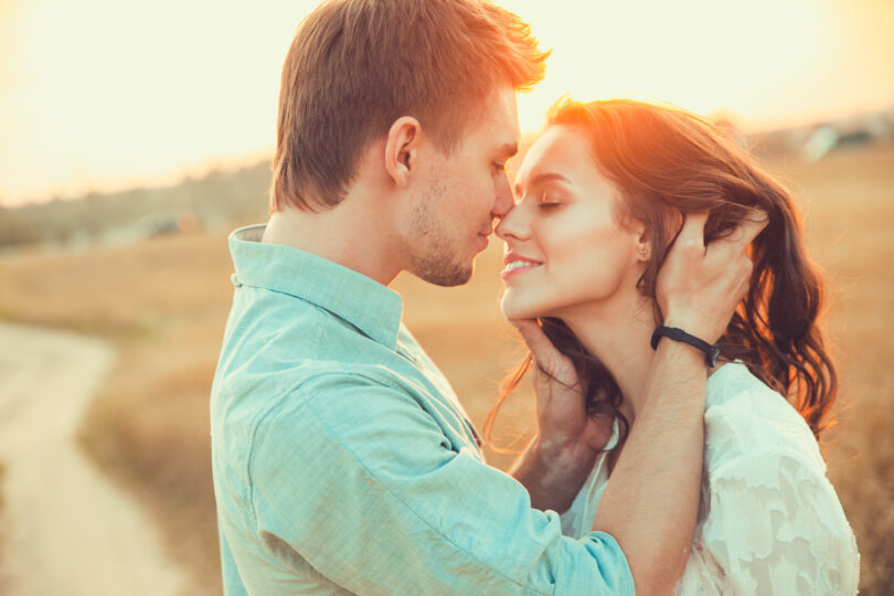 Casal em um campo aberto ensolarado. O homem segura o rosto da mulher bem próximo ao seu e ambos sorriem.