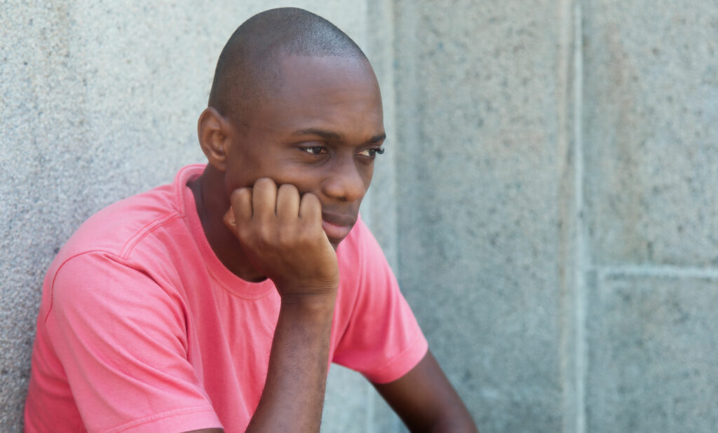 Homem negro, jovem, triste, sentado no chão, encostado em uma parede de cinza.