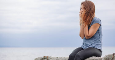 Mulher com mão no rosto e cabelo ao vento sentada em uma pedra com mar ao fundo