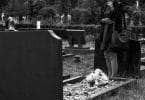 Mulher em pé em frente a um túmulo em um cemitério, demonstrando luto e tristeza.. Sua mão está cobrindo o rosto e há um buquê de flores sobre o túmulo.