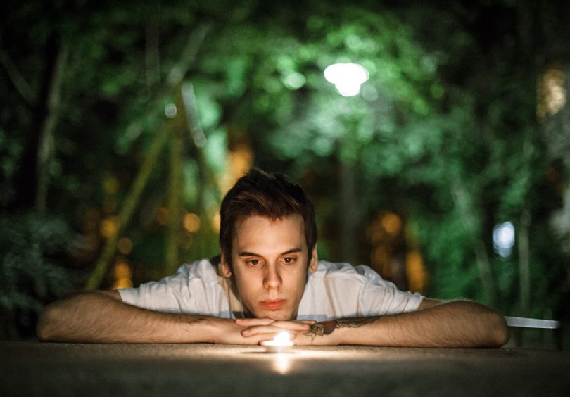 Homem branco, apoiando-se em uma mesa, observando uma pedra luminosa, com plantas ao fundo.