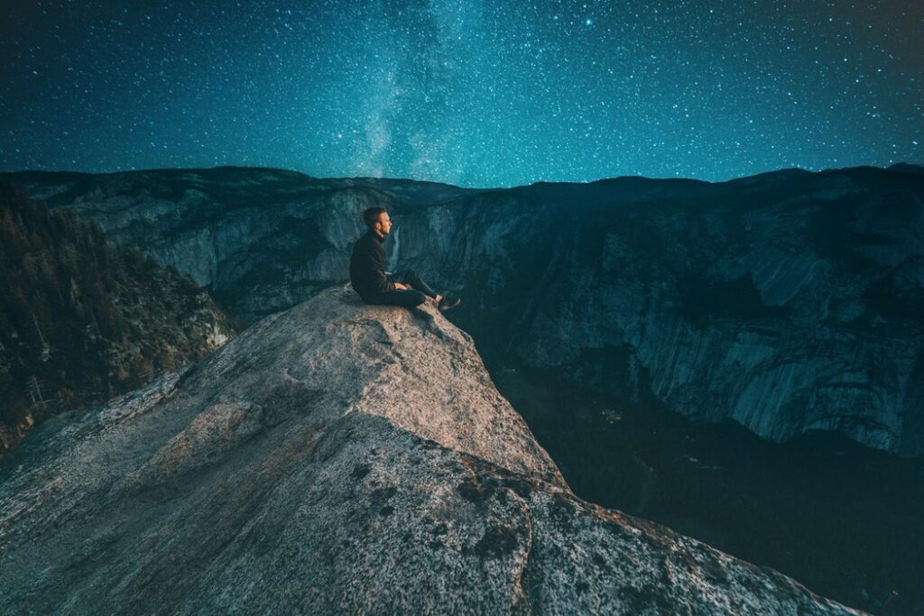 Homem sentado na beira de um penhasco, observando o céu azul da noite estrelada.