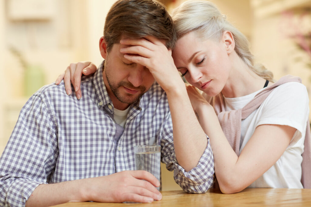 Homem e mulher, ambos brancos e jovens, sentados em uma. Ambos com expressão de preocupados. A mulher está abraçando o homem.