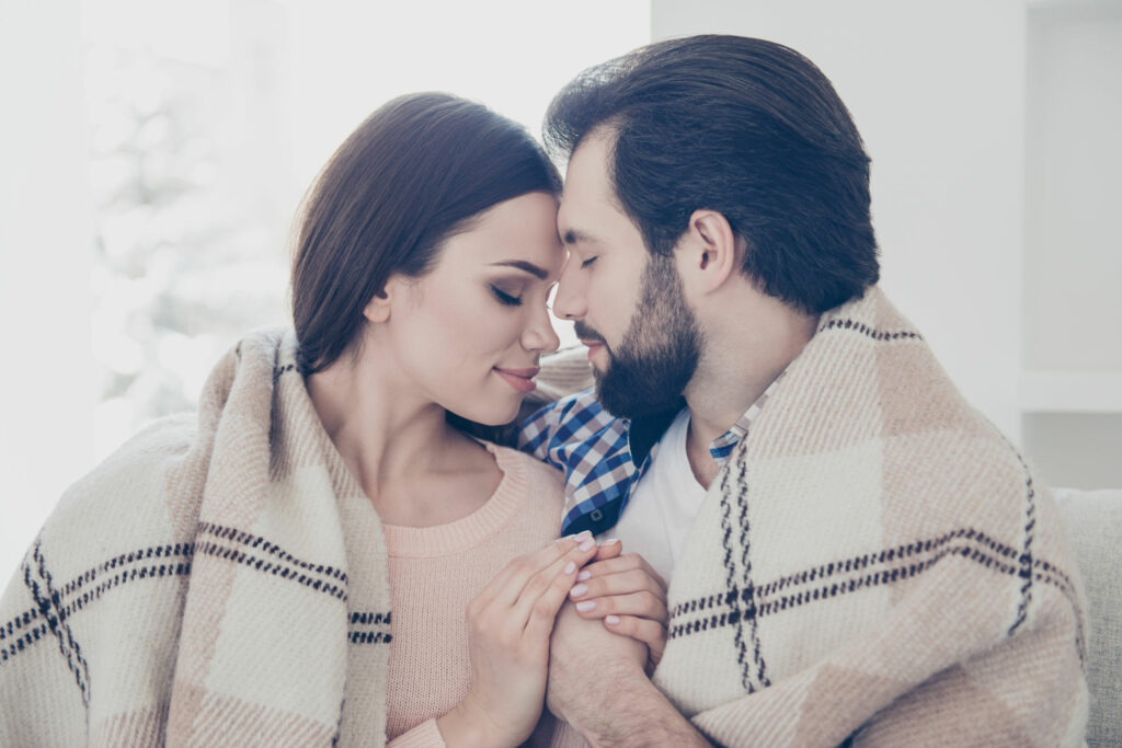 Casal sentado em um sofá, abraçado, se cobrindo com um cobertor, encostando seus rostos um no outro.