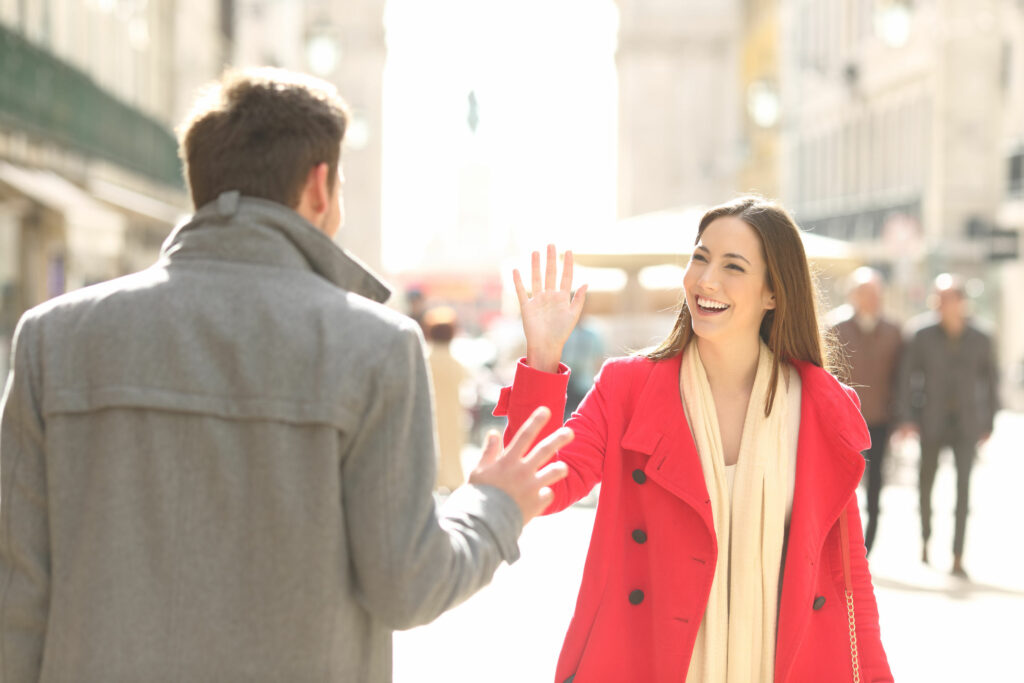 Mulher branca, jovem, sorridente, andando na rua, cumprimentando de longe um homem.