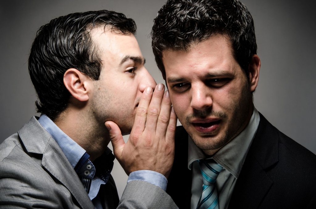 Dois homens brancos, vestindo roupas sociais, conversando, um cochichando no ouvido do outro, contando uma fofoca.