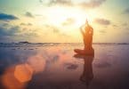 Mulher na beira da praia sentada em posição de ioga com céu ao fundo e luz do refletida