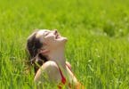 Garota feliz sentada no meio da grama alta com rosto para cima e olhos fechados iluminada pelo sol