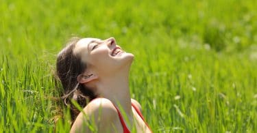 Garota feliz sentada no meio da grama alta com rosto para cima e olhos fechados iluminada pelo sol