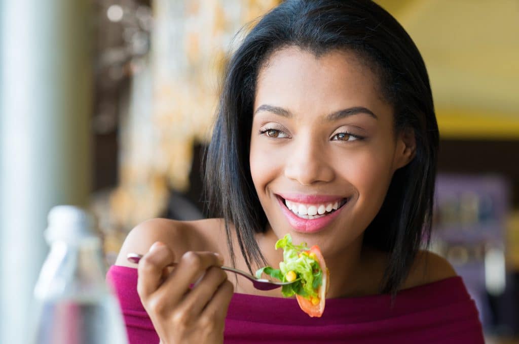 Mulher negra, jovem, sorridente, comendo uma salada.
