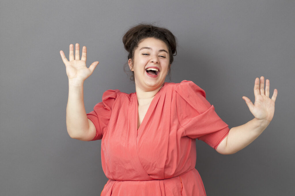 Mulher branca, gorda, vestindo um vestido salmão, sorridente, em frente à uma parede cinza