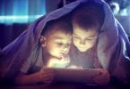 Duas crianças mexendo em um tablet embaixo das cobertas.