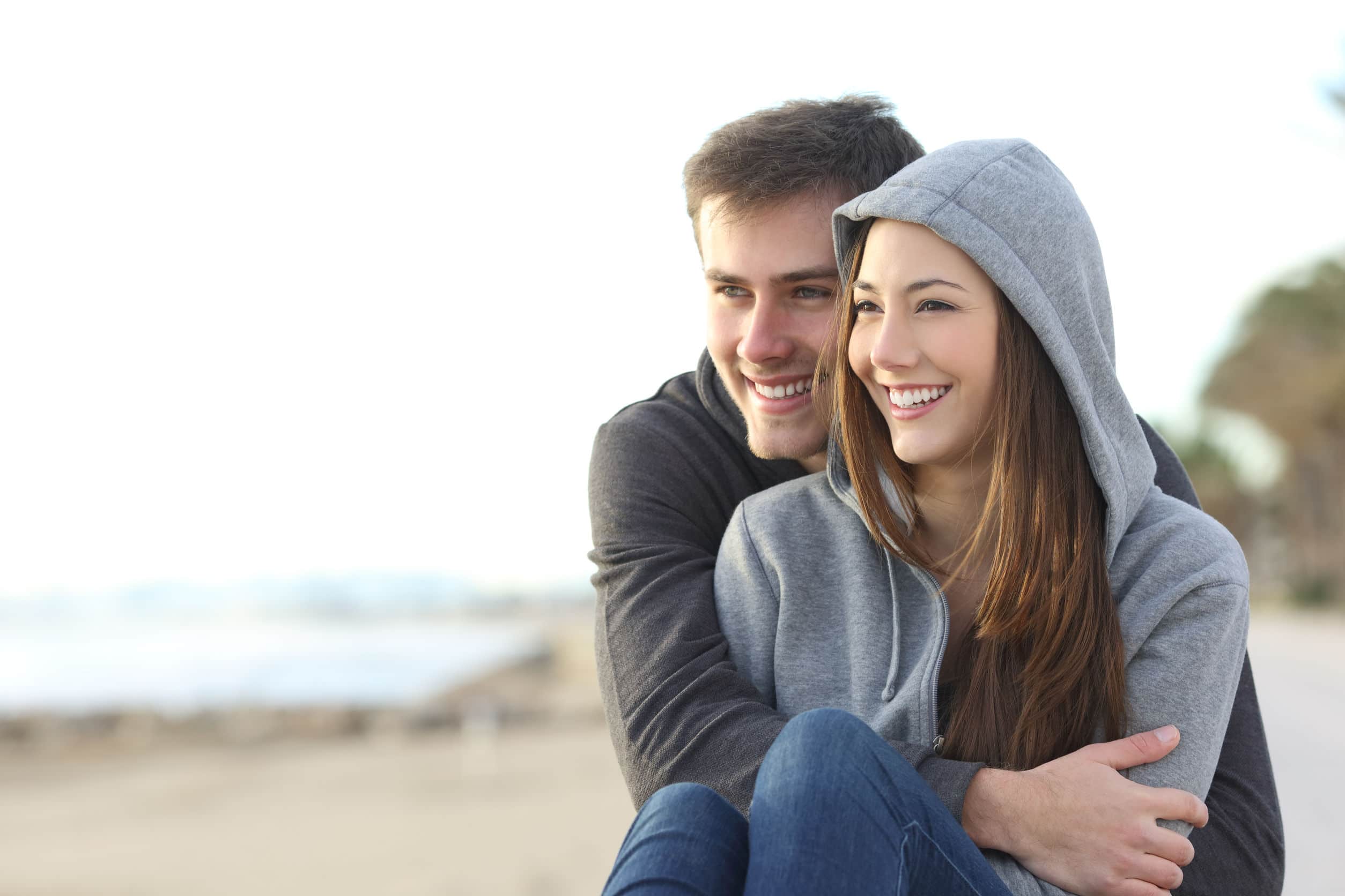 Dois adolescentes, homem e mulher, ambos brancos, vestindo blusas de moletom cinza e calça jeans, abraçados, sentados na areia de uma praia