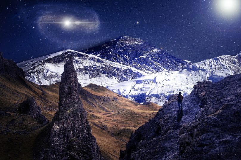 Homem no pico de montanhas nevadas, observando o brilho de um astro e das estrelas no céu.