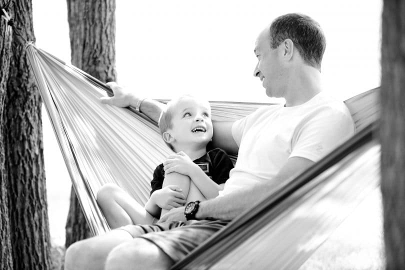Pai e filho sentados em uma rede. O menino olha para o pai e ambos sorriem. A imagem é em preto e branco.