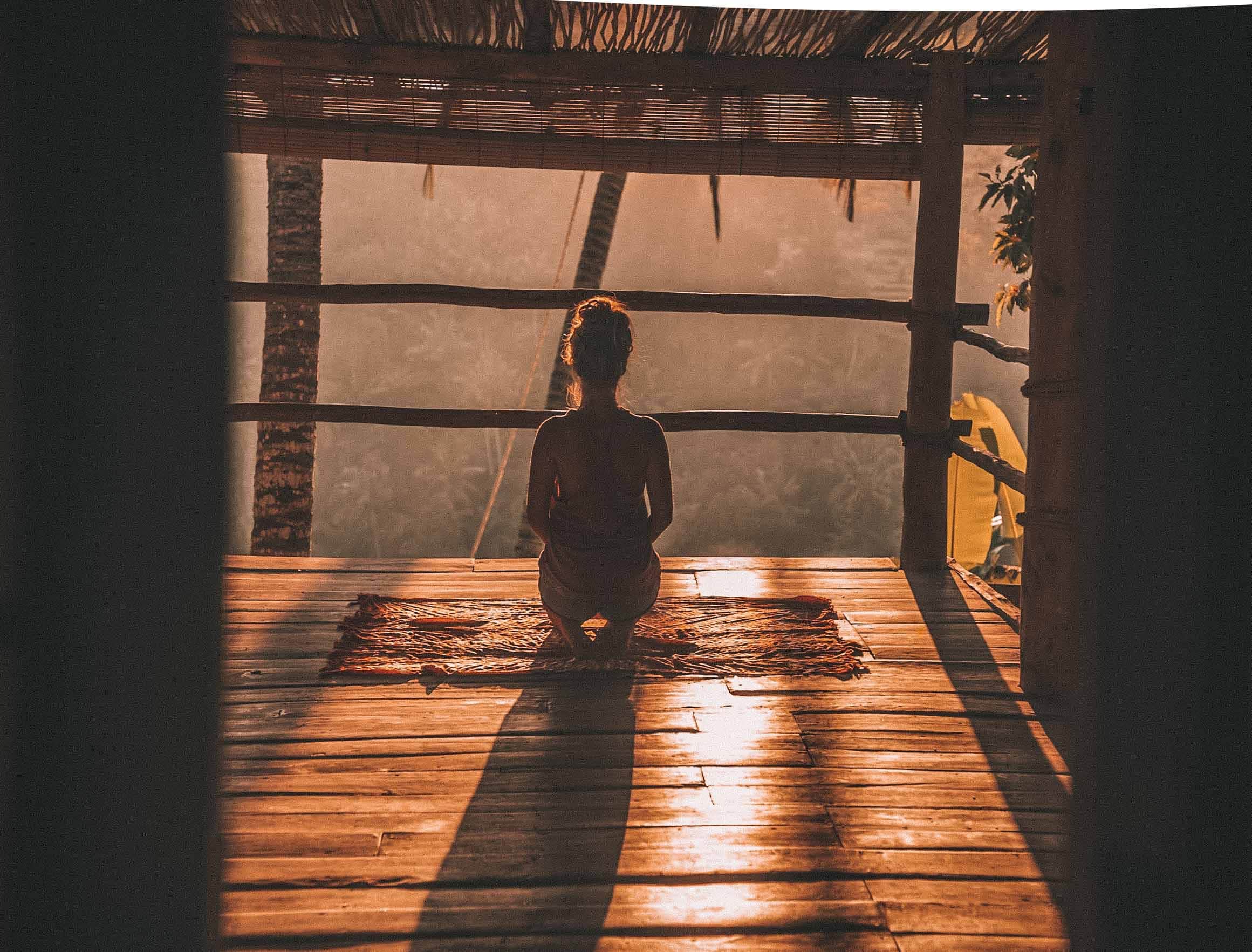 Mulher em varanda de casa de madeira de costas de frente para o sol meditando