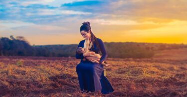 Mãe amamentando seu filho em um campo aberto. Ela está vestindo um vestido roxo e ao fundo há uma linda paisagem de entardecer.