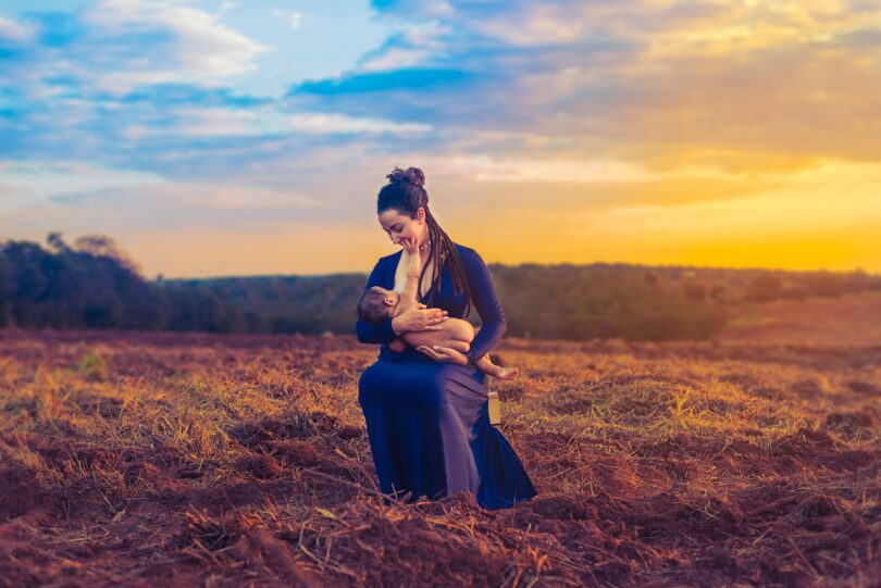 Mãe amamentando seu filho em um campo aberto. Ela está vestindo um vestido roxo e ao fundo há uma linda paisagem de entardecer.