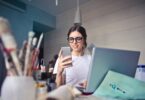 Mulher sorridente, sentada em uma mesa de escritório, olhando para o seu celular, com seu notebook aberto e funcionando.