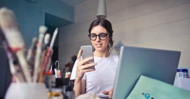Mulher sorridente, sentada em uma mesa de escritório, olhando para o seu celular, com seu notebook aberto e funcionando.