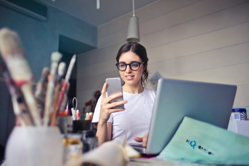 Mulher sorridente, sentada em uma mesa de escritório, olhando para o seu celular, com seu notebook aberto e funcionando.