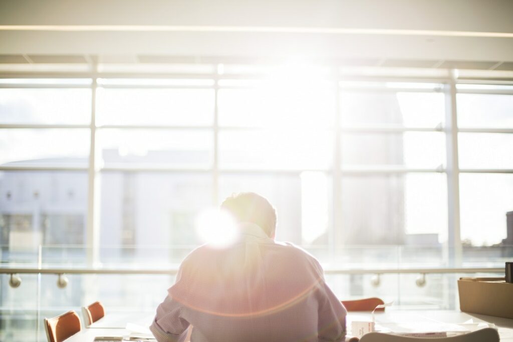 Homem vestindo roupas sociais, sentado em uma mesa de reuniões, em frente à uma janela onde o sol está batendo. 