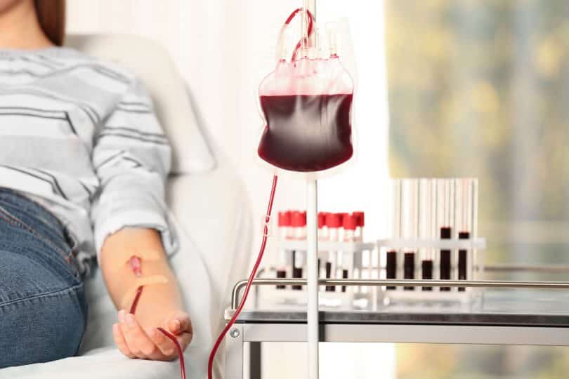 Pessoa doando sangue, ao lado de um carrinho com tubos de exames.