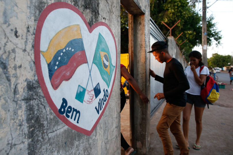 Muro com coração pintado com as bandeiras de Venezuela e Brasil pintadas escrito: Bem-vindos! E portão com pessoas entrando