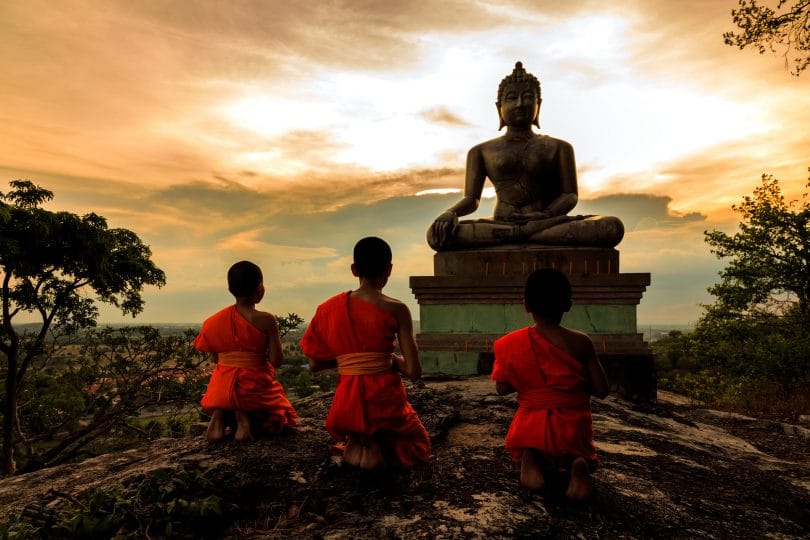 Três pequenos monges observando a estátua de um monge budista ao pôr-do-sol.