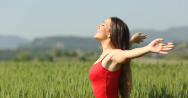 Mulher em campo verde com braços abertos sendo iluminada pelo sol