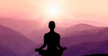 Silhueta de uma pessoa de costas meditando com o sol e montanhas ao horizonte.