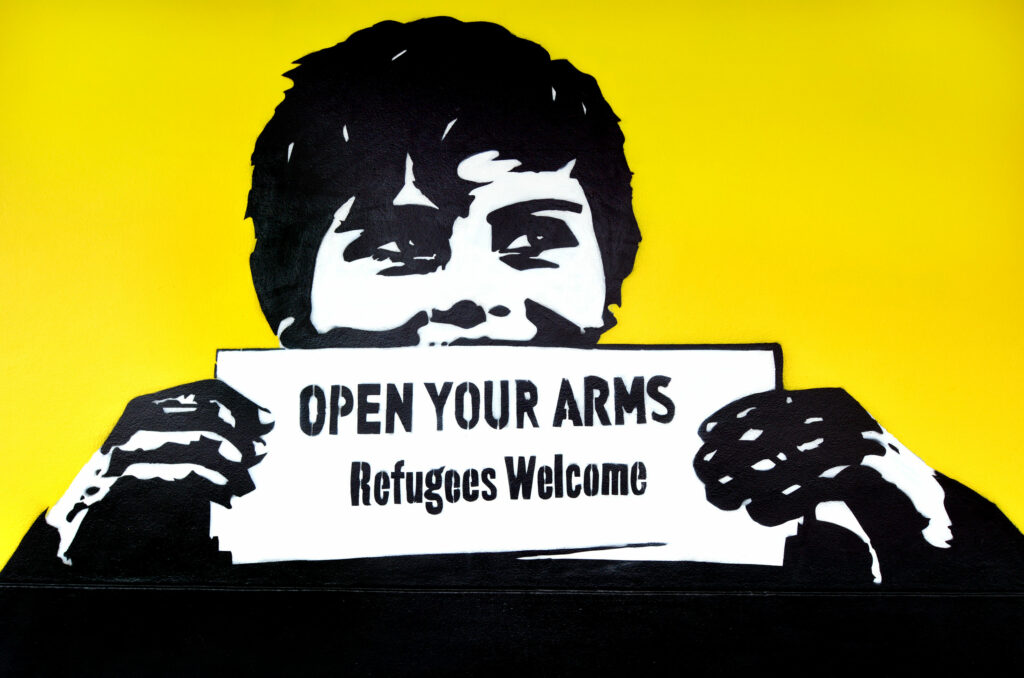 Ilustração de grafite de criança segurando cartaz escrito em inglês: abra seus braços e receba os refugiados