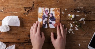 Mãos de pessoa branca segurando uma foto de casal rasgada ao meio, em cima de uma mesa de madeira, representando o término de relacionamento.