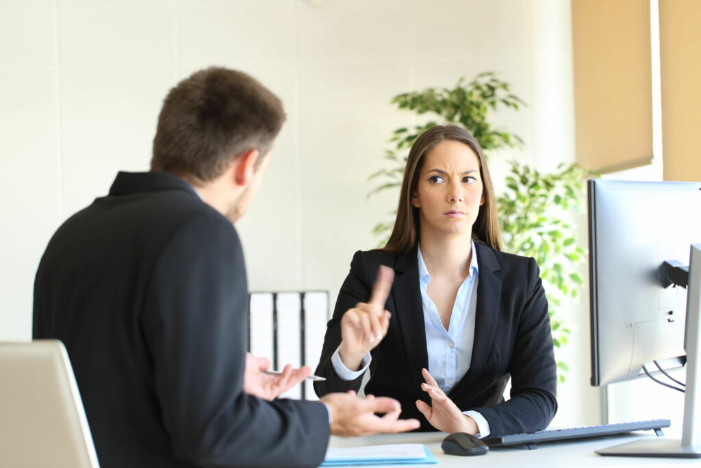 Homem e mulher, ambos brancos, vestidos de social, em um escritório. Mulher sentada atrás de uma mesa, falando não, gesticulando com a mão, para o homem que está sentado na sua frente. 