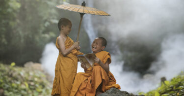 Dois pequenos monges budistas conversando e rindo. Um deles está segurando um guarda chuva e o outro lê um livro. Ambos estão sentados em uma pedra na floresta.