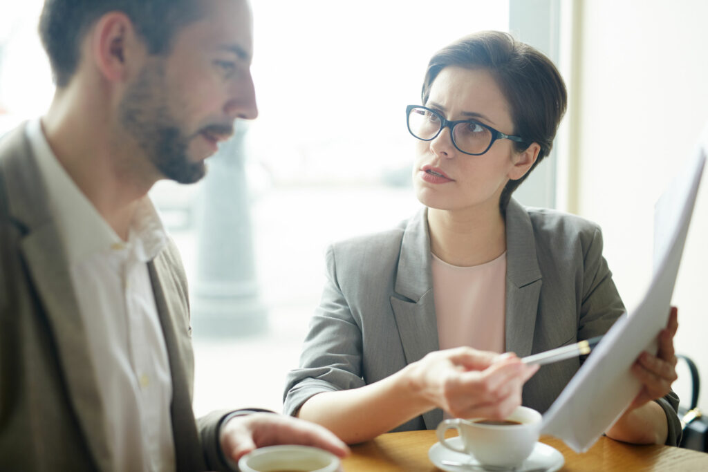 Mulher e homem, ambos vestindo roupas sociais, sentados em uma mesa, tomando café e conversando sobre um documento que está na mão da mulher