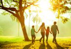 Família andando de mãos dadas em um campo, ao lado de uma árvore e com o sol brilhando ao fundo.
