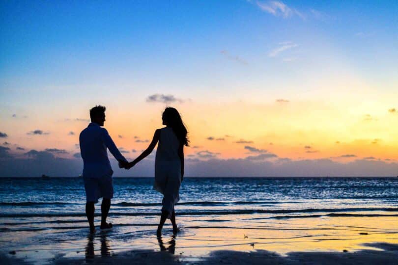 Casal andando na beira da praia com pôr do sol de fundo