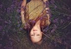 Garota deitada em grama com flores roxas sobre seu corpo de cabelo solto