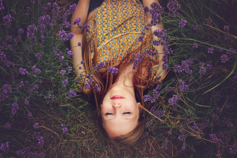 Garota deitada em grama com flores roxas sobre seu corpo de cabelo solto
