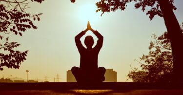 Silhueta de pessoa com mãos acima da cabeça sentada em posição de meditação com árvores e sol ao fundo