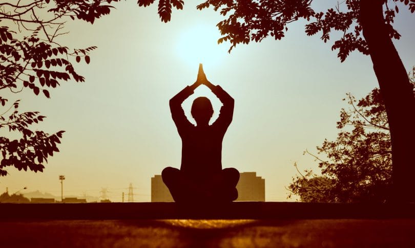 Silhueta de pessoa com mãos acima da cabeça sentada em posição de meditação com árvores e sol ao fundo