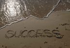 "Success" escrito na areia com água do do mar ao lado