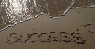 "Success" escrito na areia com água do do mar ao lado
