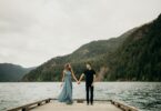 Casal em pé de mãos dadas em cima de ponte com mar e montanha de fundo