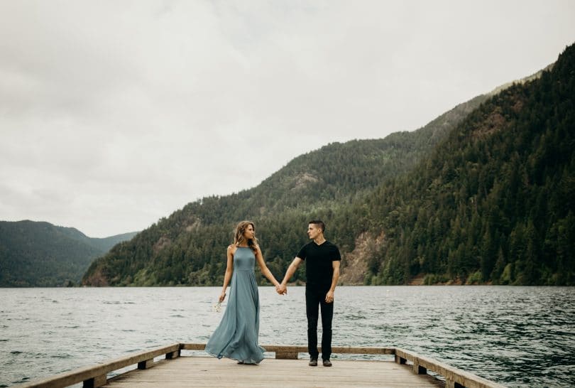 Casal em pé de mãos dadas em cima de ponte com mar e montanha de fundo