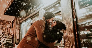 Casal se beijando com a neve caindo em cima deles.